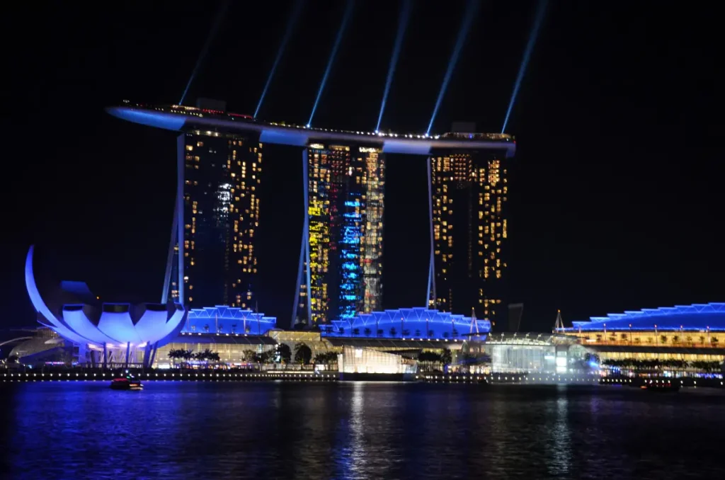 A nighttime view of the Marina Bay Sands in Singapore, illuminated by a vibrant light show with blue and white beams stretching into the sky. The ArtScience Museum's lotus-shaped structure and surrounding waterfront buildings are lit in striking blue, reflecting on the calm water.