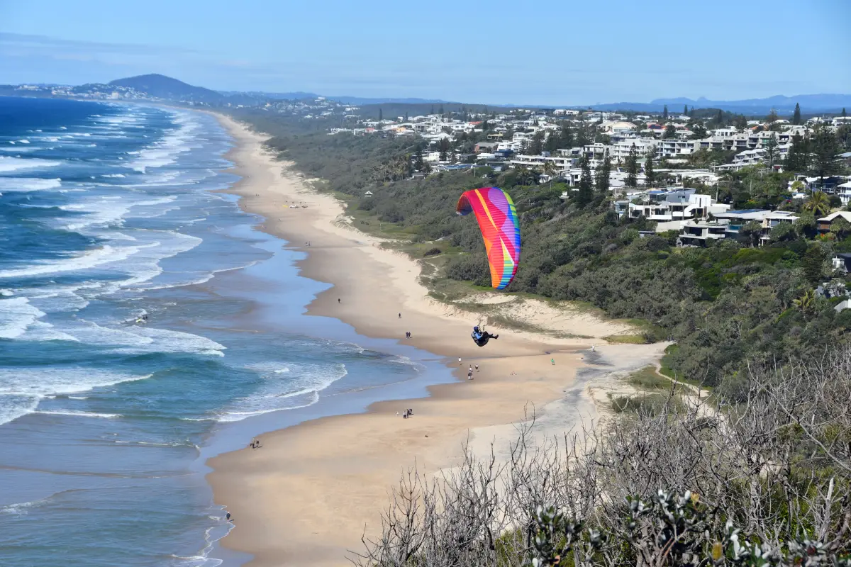 paraglider over long strecth of sandy beach with houses backing onto beach