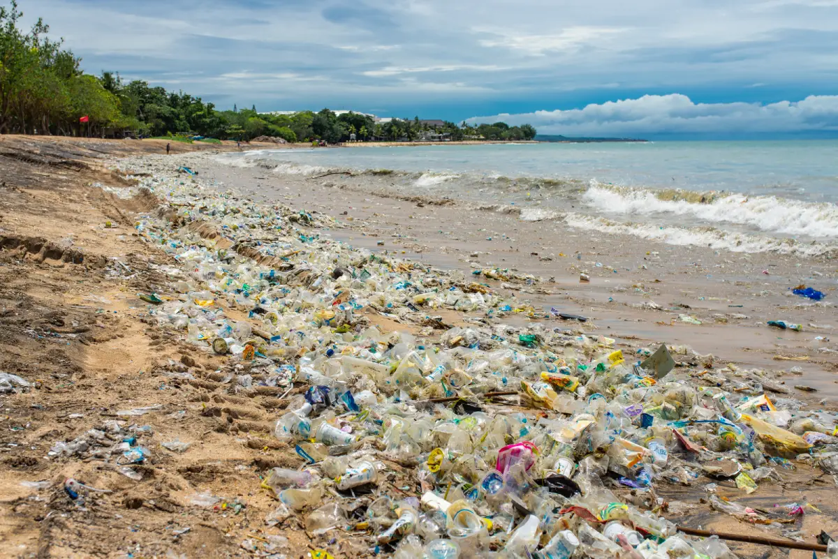 A once-beautiful beach is now covered in plastic waste, with countless bottles, wrappers, and other debris littering the sandy shore. The ocean waves continue to wash more trash onto the beach, highlighting the severe issue of marine pollution. In the background, lush greenery and distant buildings contrast sharply with the environmental damage in the foreground