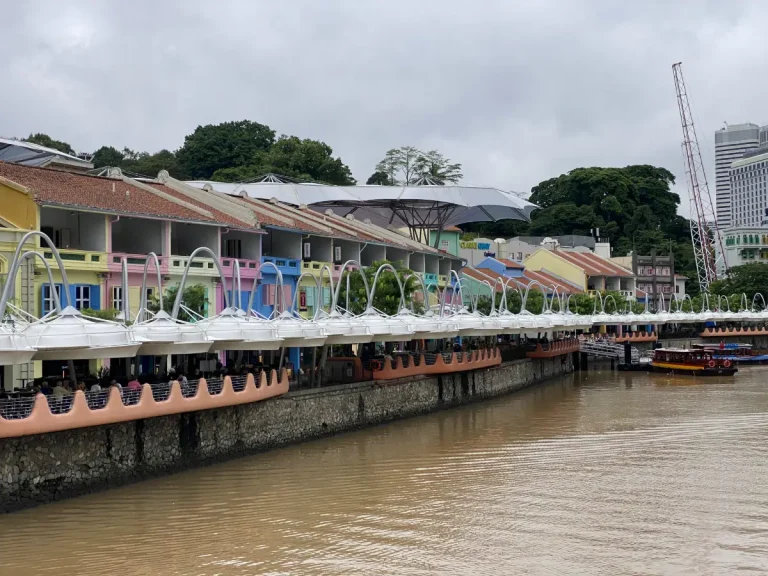 This image showcases Clarke Quay in Singapore, a riverside dining and entertainment hub lined with colorful shophouses. The waterfront promenade features a modern canopy with white, curved structures covering the outdoor seating areas. Traditional bumboats are seen on the Singapore River, adding to the lively atmosphere of this popular tourist destination.