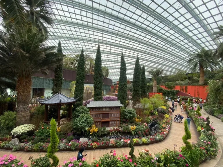 A vibrant floral display inside the Flower Dome at Gardens by the Bay, featuring a traditional Asian-style pavilion and a miniature wooden building surrounded by colorful flowers and neatly trimmed trees. The glass ceiling arches overhead, allowing natural light to illuminate the lush greenery and winding pathways filled with visitors