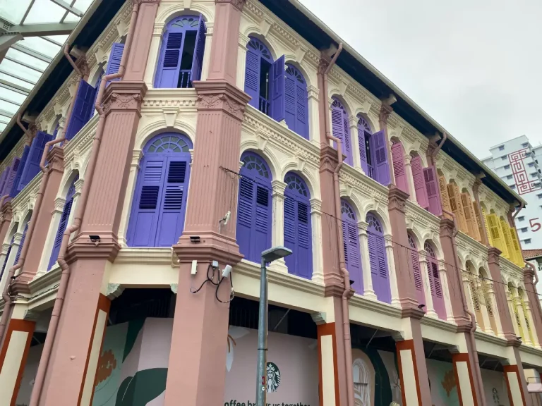 This image showcases a beautifully preserved colonial-style shophouse in Singapore's Chinatown, featuring pastel-colored shutters in shades of purple, pink, and yellow. The intricate architectural details, including arched windows and ornate columns, highlight the building's historical charm. The lower level appears to house a commercial space, adding to the vibrant and dynamic streetscape of the area.