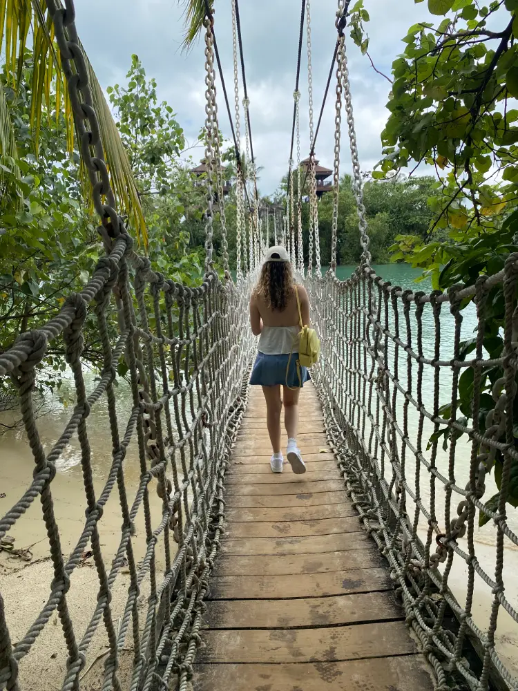 A woman walks across a rope suspension bridge over turquoise water surrounded by lush greenery on Sentosa Island, Singapore — a tropical escape blending adventure and natural beauty